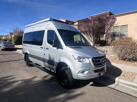 A silver Mercedes-Benz Sprinter camper van for RV rental, parked on a sunny residential street.