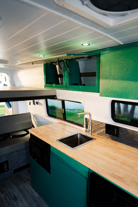 This RV rental features a modern interior with a wood countertop kitchenette, sink, green cabinets, and a bed. Natural light from windows fills the space.