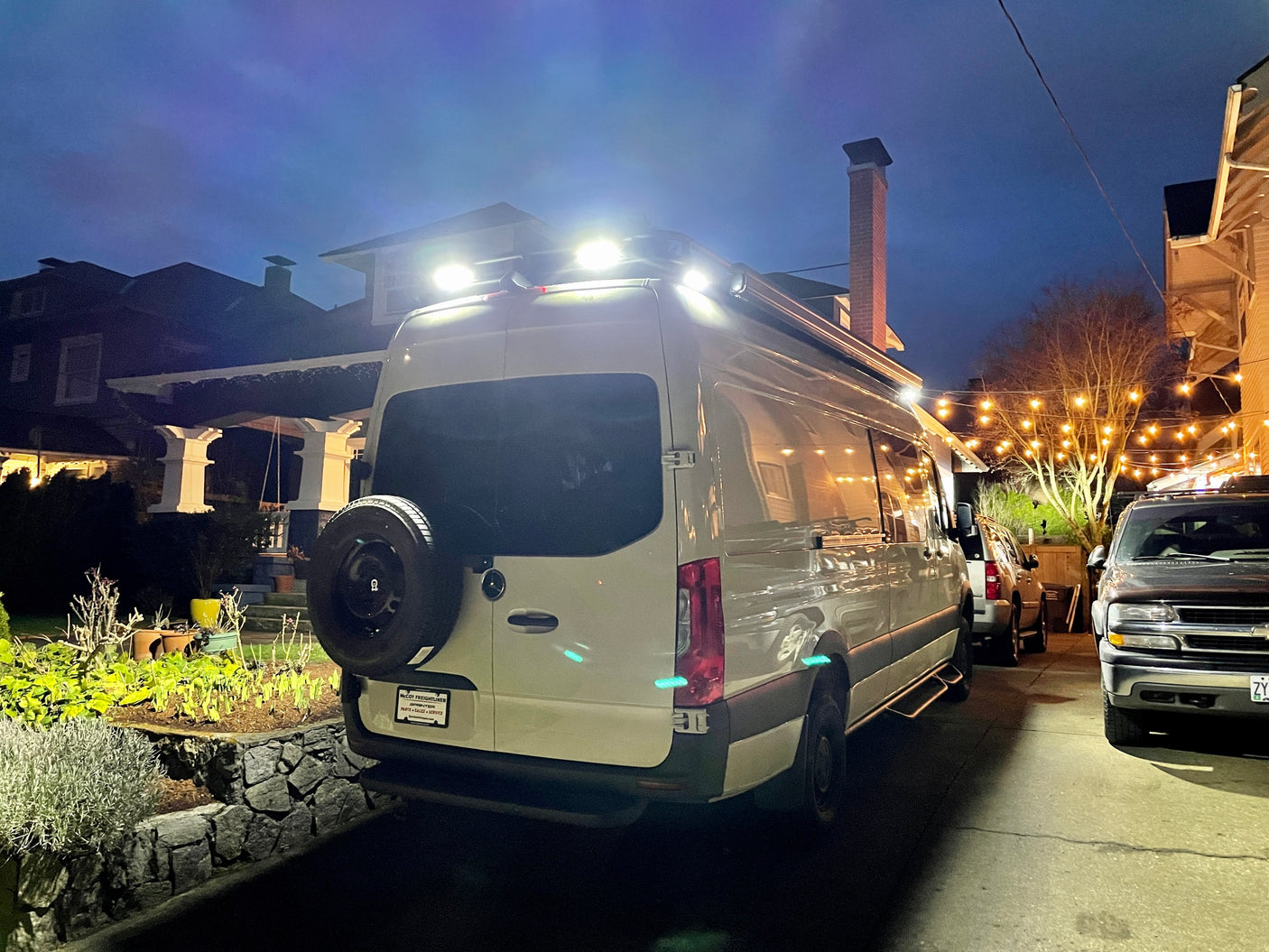 White Sprinter rv rental van with bright rooftop lights and rear spare tire, parked at night in a residential setting.