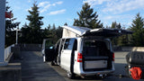 Silver pop-top RV rental camper van with open doors and a cargo box. Parked in a concrete lot amidst green trees.
