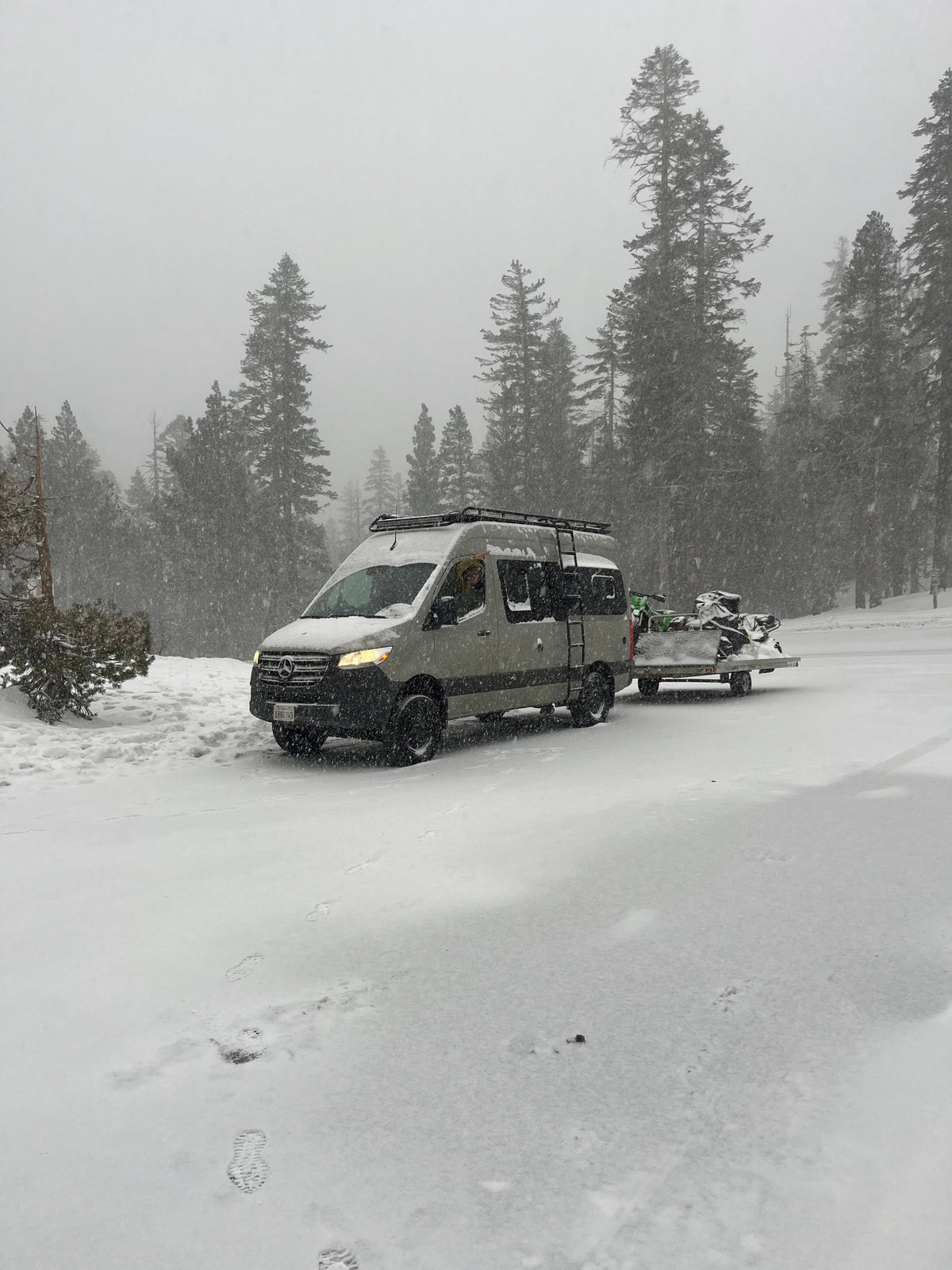 A gray Mercedes-Benz RV rental with a roof rack is towing snow-covered snowmobiles through a snowy forest. — photo 6