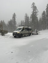 A gray Mercedes-Benz RV rental with a roof rack is towing snow-covered snowmobiles through a snowy forest.