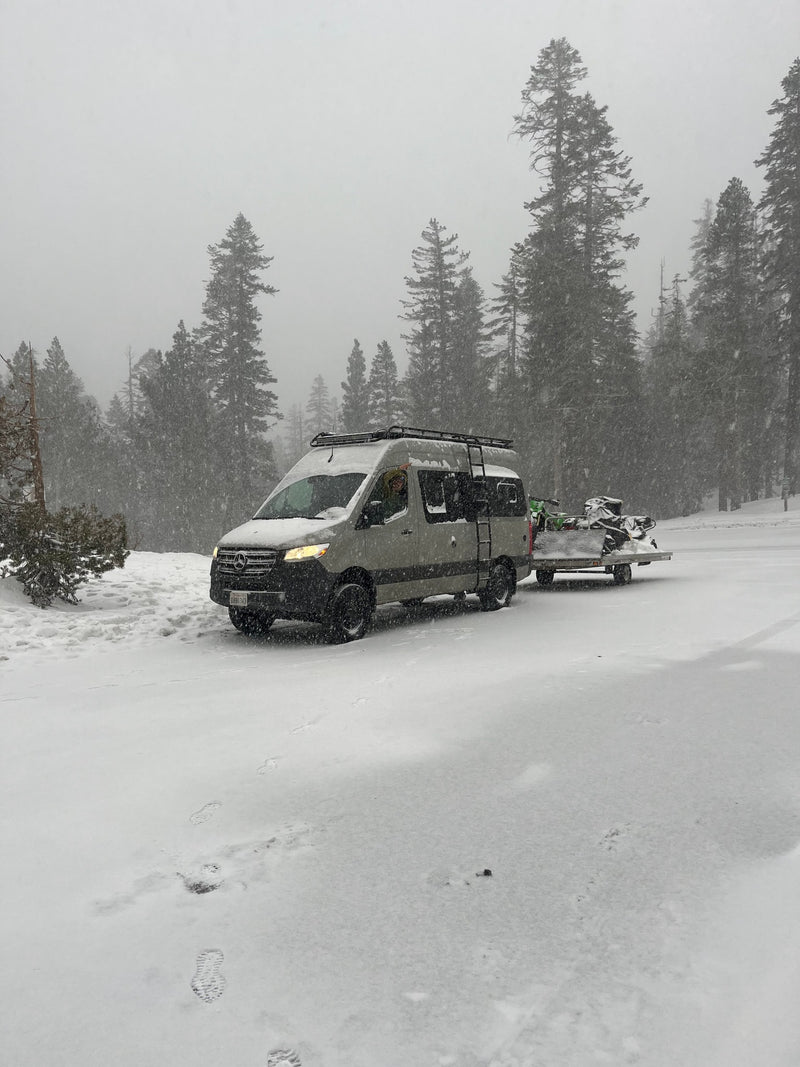 A gray Mercedes-Benz RV rental with a roof rack is towing snow-covered snowmobiles through a snowy forest. — photo 6