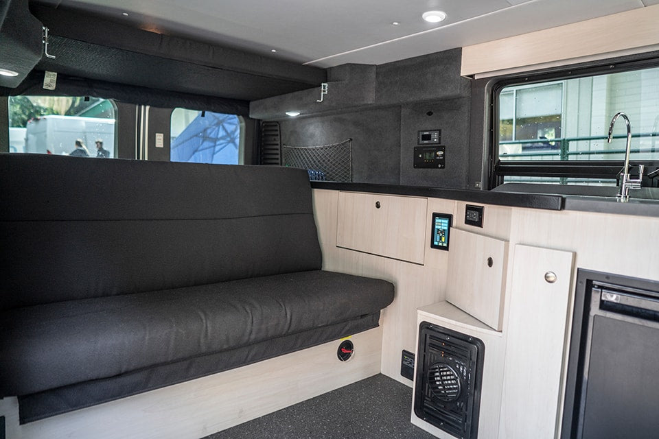 RV rental interior: a dark grey sofa, light wood cabinetry, modern kitchen with sink, and digital control panels are visible. — photo 11