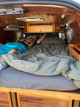 Enjoy an RV rental: Man relaxing on a gray bed inside a cozy, wood-paneled camper van interior.