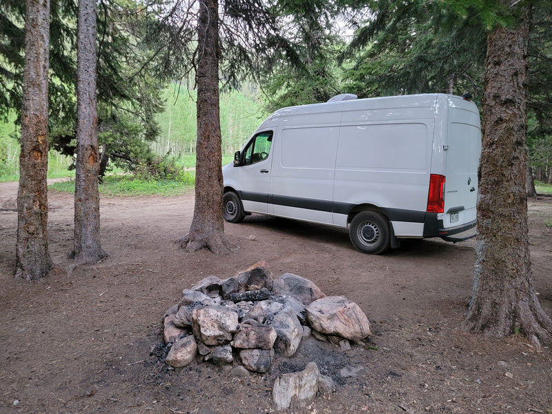 RV rental camper van parked in a forest campsite with a stone fire pit. Tall trees surround the scene. — photo 6