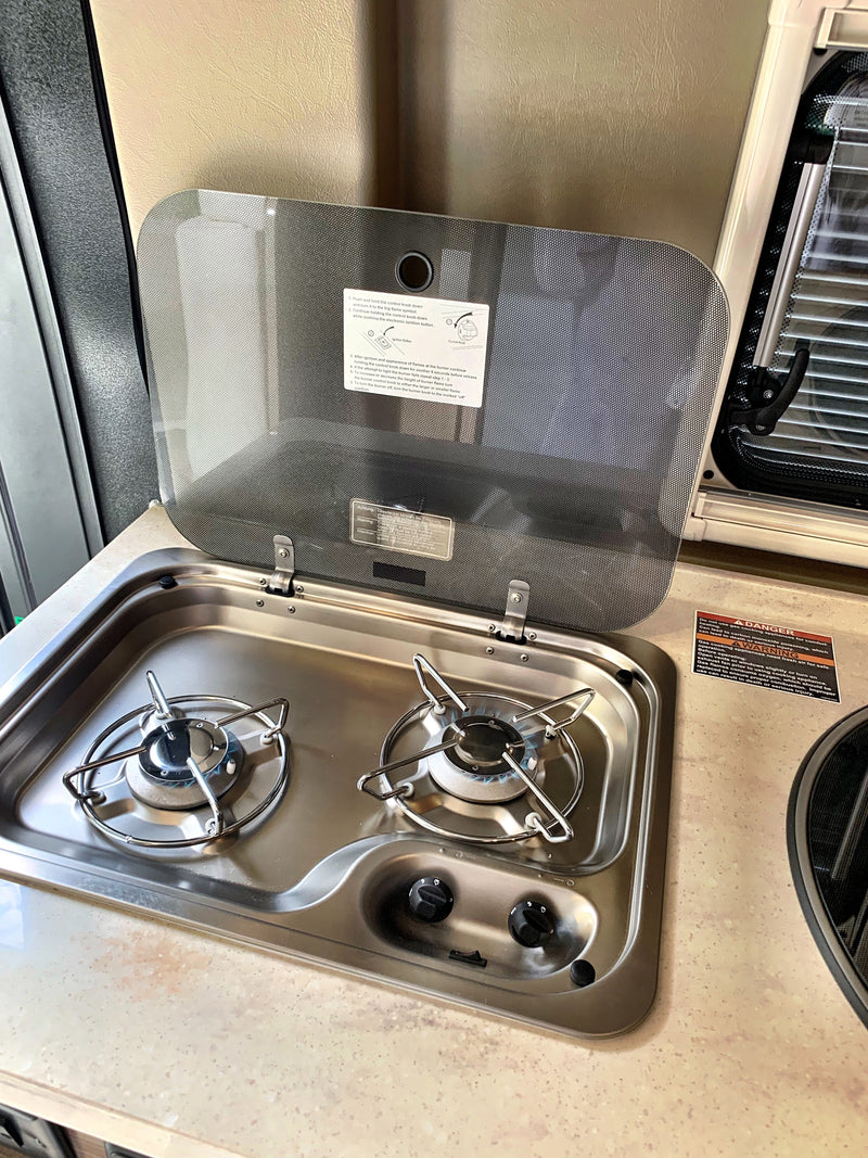 RV rental kitchen featuring a two-burner gas cooktop with an open safety lid, showing usage instructions and a lit burner. — photo 7