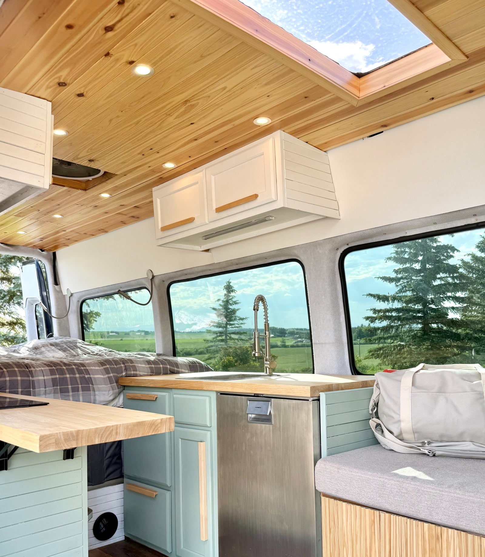 This rv rental's spacious camper van interior features a wood ceiling, skylight, light blue kitchen, and a bed with scenic views.