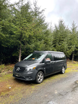 An RV rental adventure awaits! This Mercedes-Benz pop-top camper van is parked on a wet road by a dense forest.