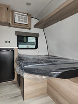Interior of an rv rental showing a compact fridge, window AC unit, and a new bed still wrapped in protective plastic.
