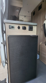RV rental: Camper interior with a built-in refrigerator, a wood panel featuring power outlets and switches, and compact storage.