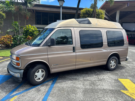 Tan Chevy 1500 Discovery high-top van, perfect for rv rental, parked by a building and tropical plants.