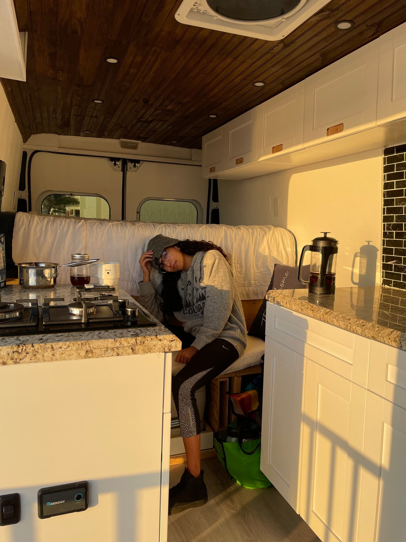 Inside a cozy RV rental: a woman relaxes in the sun-drenched kitchen area with stove, French press, and wooden ceiling.