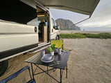 Beachside living with an **rv rental**: A camper van parked on sandy beach with a table set up, facing a mountain and ocean.