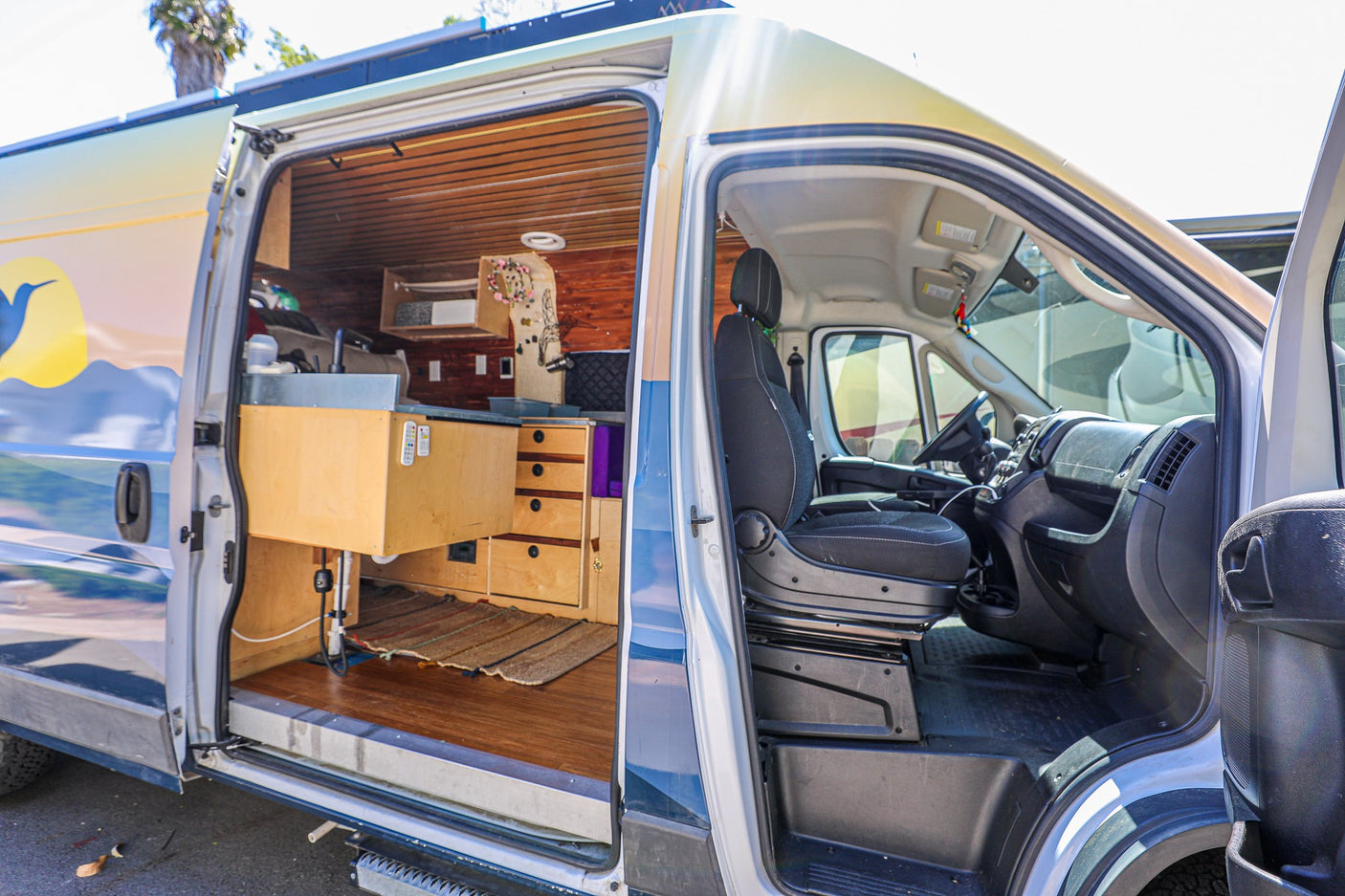 Open RV rental: a custom camper van's interior with wood paneling, counter, and driver's cabin visible, plus a mountain mural on its exterior.