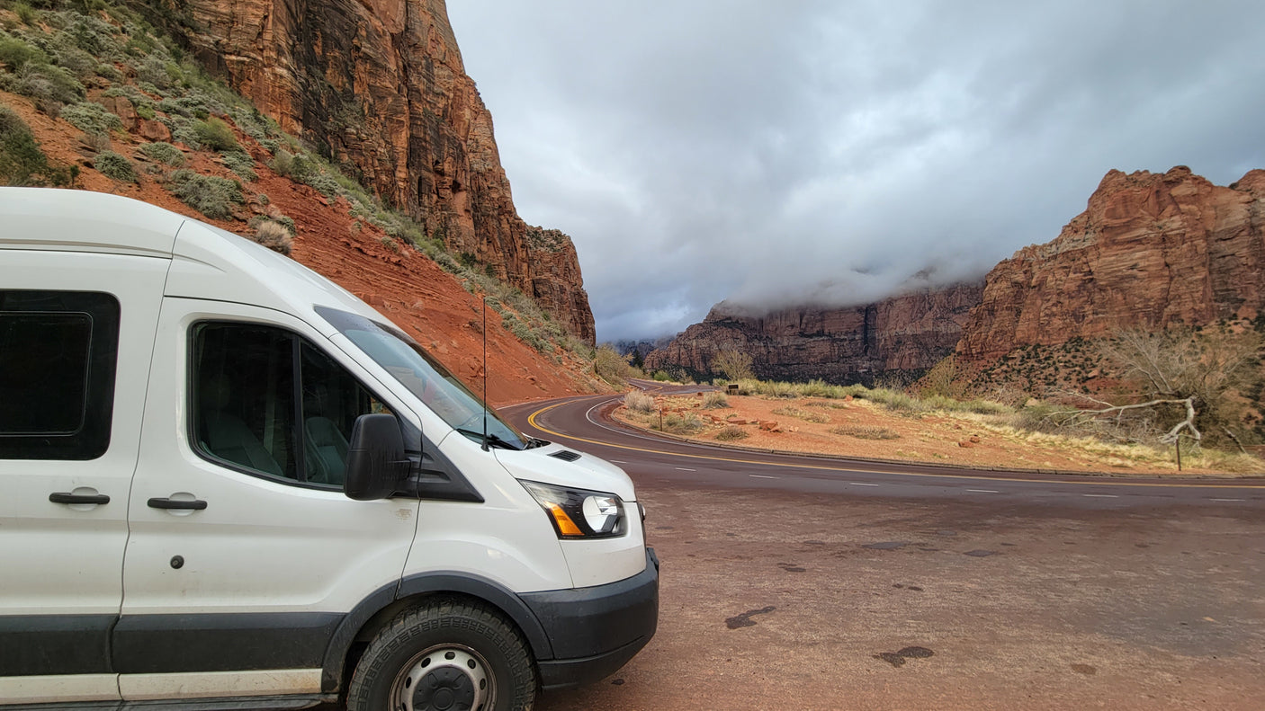 A white camper van (rv rental) on a scenic mountain road, majestic red rock landscape under cloudy sky. Ideal for a road trip.