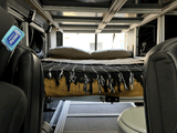 Cozy RV rental interior: A view of the elevated bed with pillows and a blanket, showing a modular frame, front seat, and toilet.