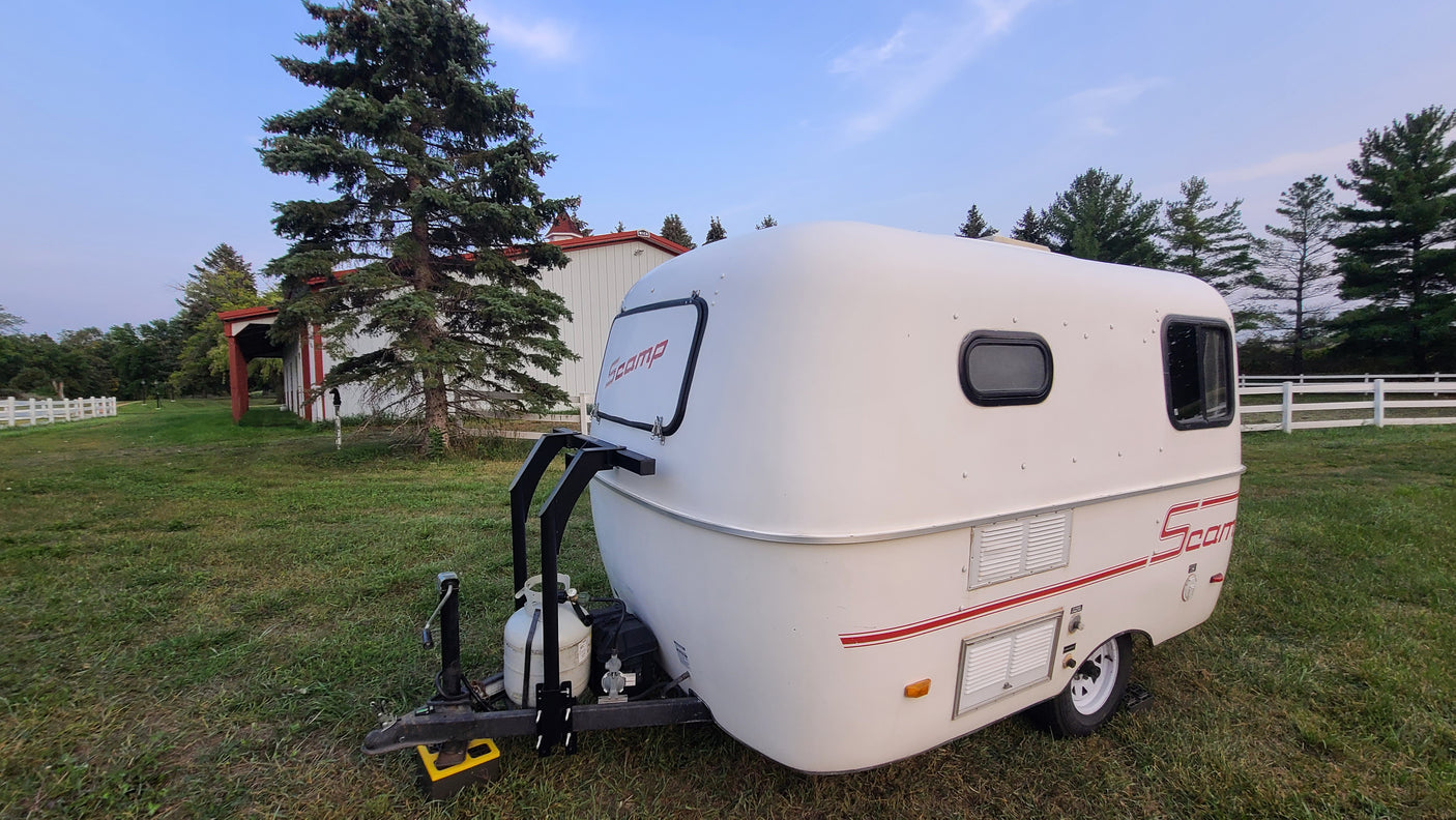 This compact white Scamp travel trailer (RV rental) is parked on grass, with a barn and pine trees in the background.