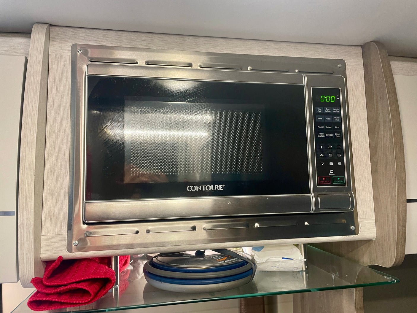 Stainless steel built-in Contoure microwave in an RV rental kitchen, with wood-grain cabinetry & a glass shelf.