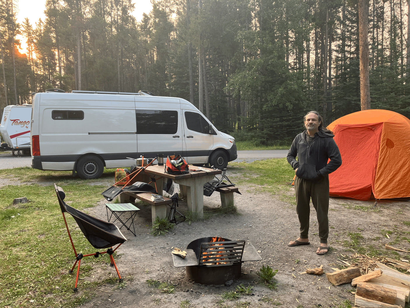 Man smiles at a peaceful campsite with a white RV rental, orange tent, and crackling campfire under tall trees. Perfect outdoor adventure.