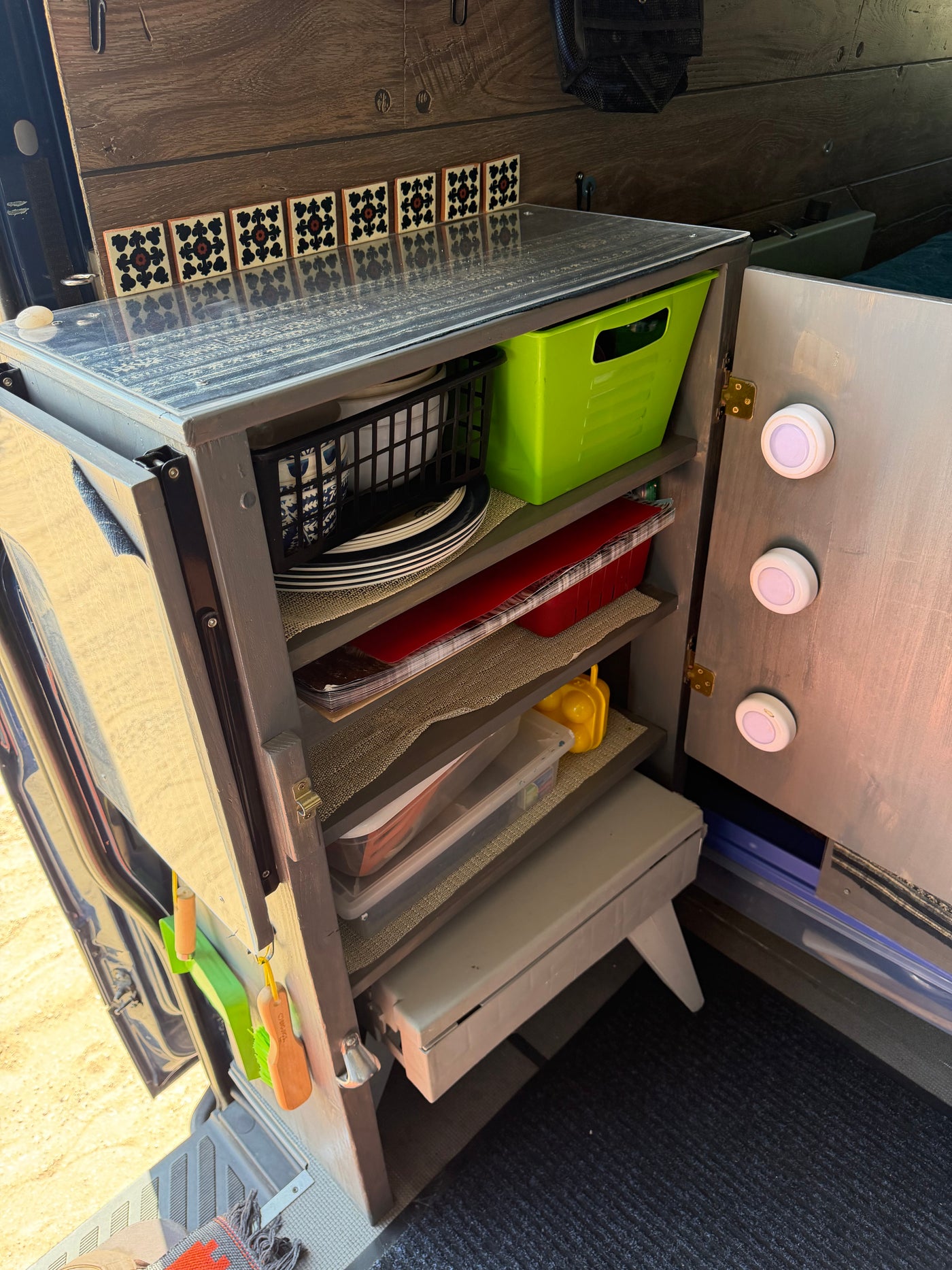 An open, organized kitchen cabinet in an rv rental, showing shelves with dishes, bins, decorative tiles, and door lights. — photo 10