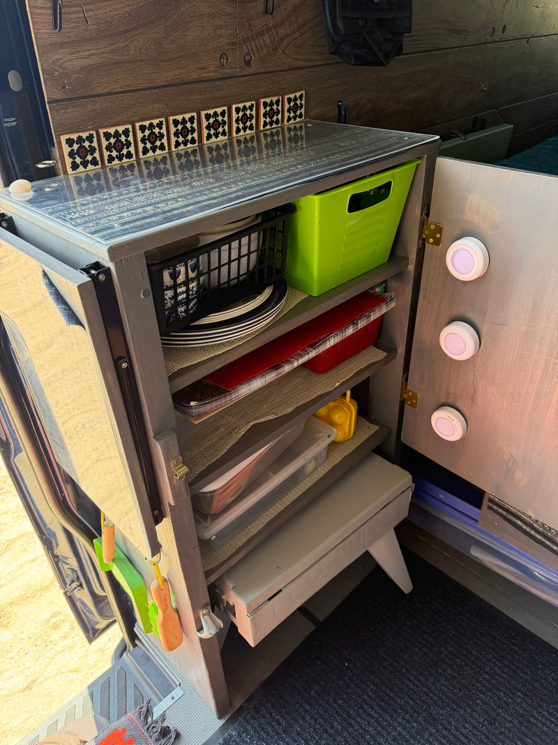 An open, organized kitchen cabinet in an rv rental, showing shelves with dishes, bins, decorative tiles, and door lights. — photo 10