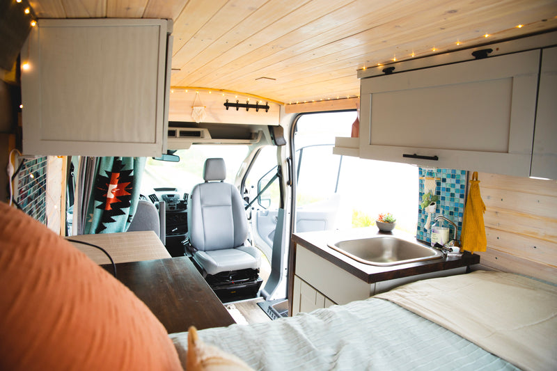 RV rental interior: A cozy, modern converted van with wood-paneled ceiling, compact kitchen, bed, and driver's cab. — photo 8
