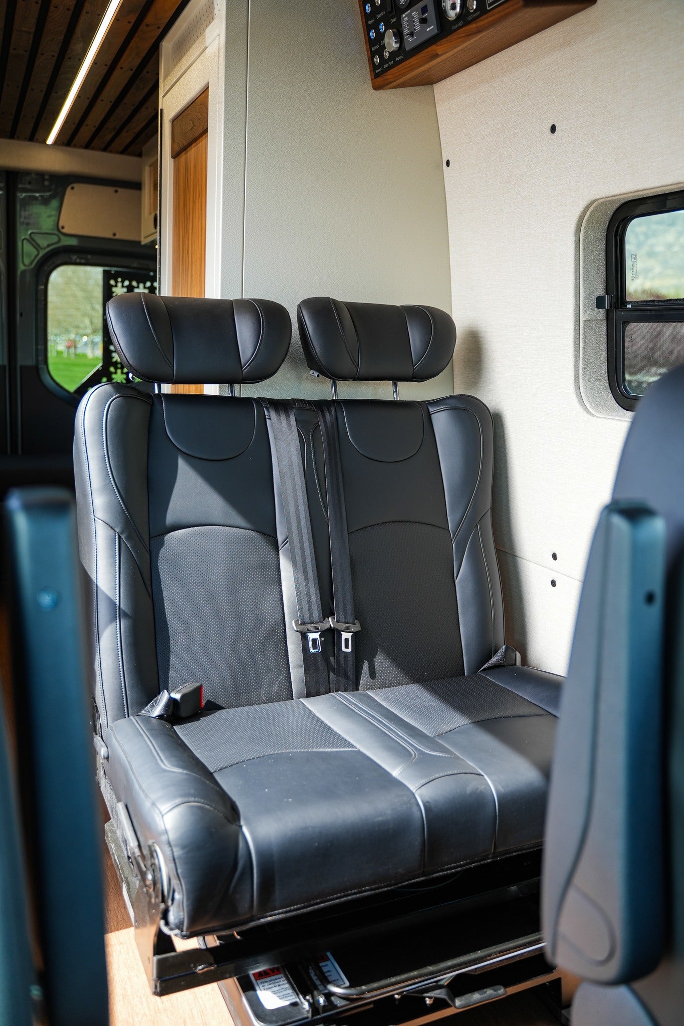 Inside an RV rental: a comfortable black leather two-person bench seat with seatbelts, wood ceiling, and control panel detail.
