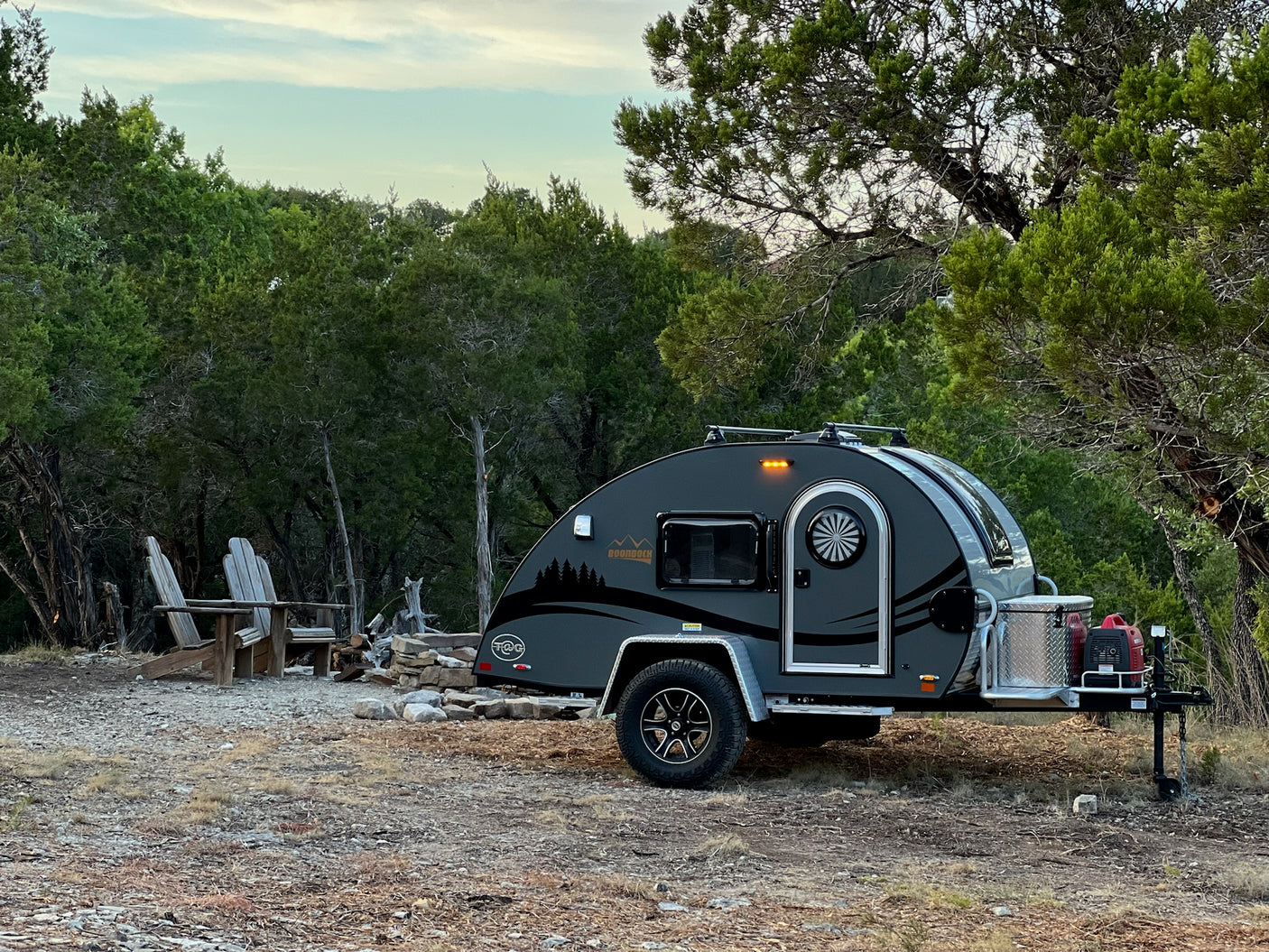 This gray teardrop RV rental is parked at a wooded campsite with two Adirondack chairs and a stone fire pit.