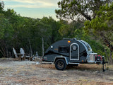 This gray teardrop RV rental is parked at a wooded campsite with two Adirondack chairs and a stone fire pit.
