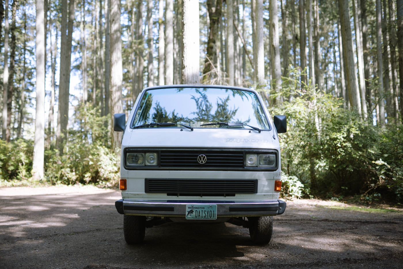  on a sunny beach. A man leans out the driver's window. Tire tracks on wet sand.|White 4WD VW Vanagon RV rental with roof rack — photo 3