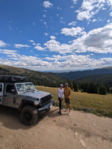 Enjoying the scenic views! A silver Jeep Wrangler **RV rental** with a rooftop tent on a mountain road, with a couple and vast landscapes.