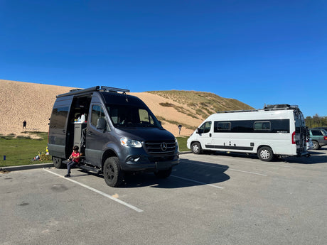 Experience van life! Two custom camper vans, resembling popular RV rental options, parked by sandy dunes. A child sits nearby.