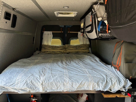 RV rental converted van interior: large bed, patterned pillows, ceiling vent, hanging storage, & wall controls.