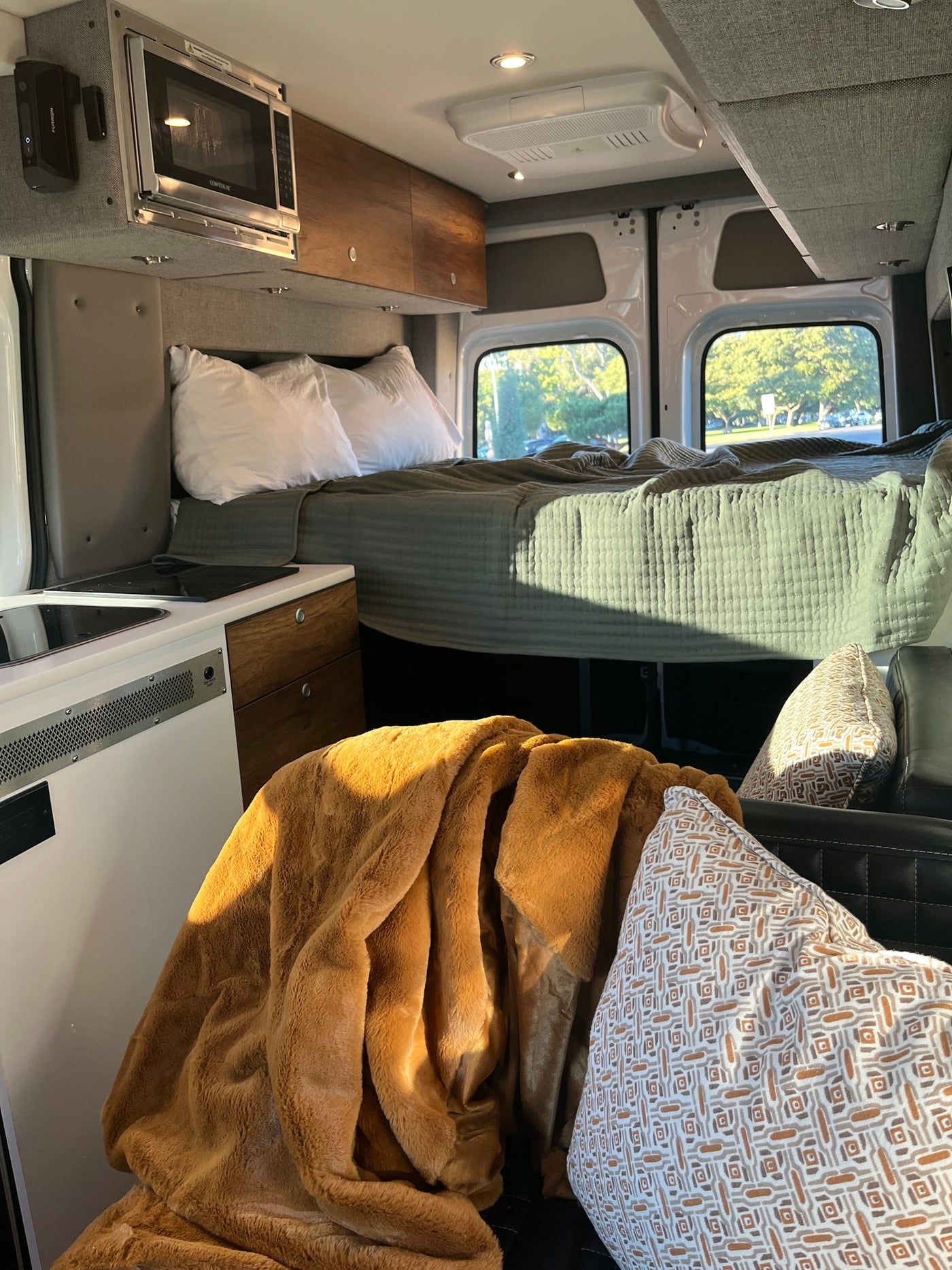 Cozy RV rental interior with a made bed, kitchen counter, overhead microwave, and rear windows showing trees outside. — photo 13