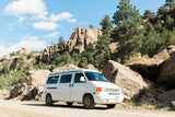 White VW RV rental van on dirt road. Scenic rocky mountains & green pines under blue sky. Ideal for nature camping & travel.