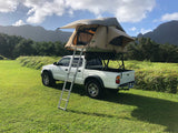 White pickup truck with a deployed tan rooftop tent & ladder on green grass, ready for your RV rental mountain adventure.