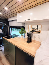 Bright RV rental interior featuring a compact kitchen with wood countertops, induction cooktop, and coffee maker, plus a bed.