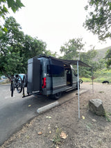 Gray Sandy Vans RV rental parked with awning out, bikes on back, ready for adventure. Dog nearby.