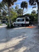 White camper van (RV rental) set up at a sandy campsite under palm trees, with an awning extended. Ideal for outdoor adventures.