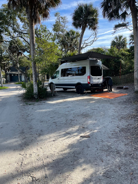 White camper van (RV rental) set up at a sandy campsite under palm trees, with an awning extended. Ideal for outdoor adventures.