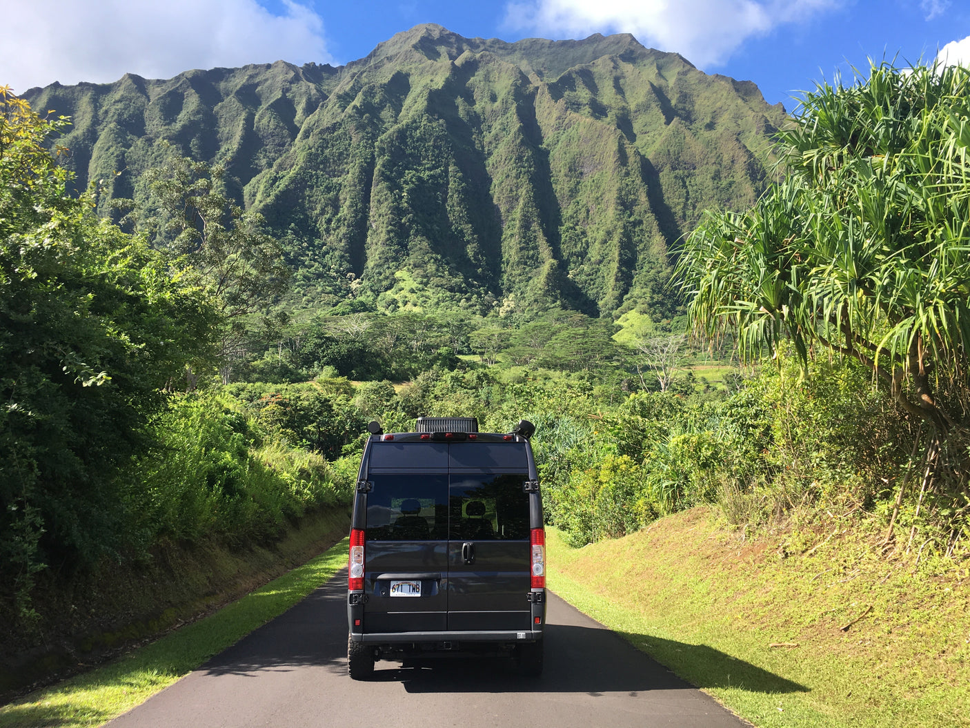 A dark RV rental van on a paved Hawaiian road, facing a majestic, verdant mountain range under a clear blue sky, amidst lush foliage.