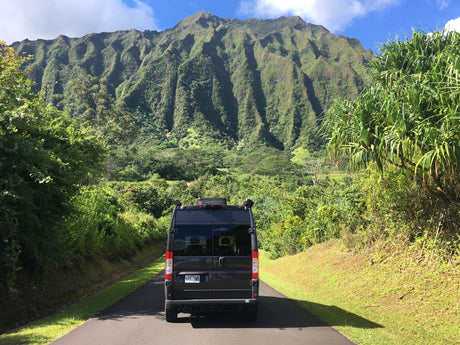 A dark RV rental van on a paved Hawaiian road, facing a majestic, verdant mountain range under a clear blue sky, amidst lush foliage.