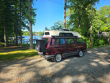 This burgundy VW camper van, ideal for RV rental adventures, is parked on a gravel path by a serene lake with docks and lush trees.