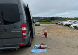 A white dog in an orange shirt patiently sits next to a grey RV rental. Gear is on the ground, cars parked in a dirt lot.