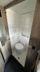 An RV rental's compact wet bath showing a white toilet, shower pan, and wall-mounted storage rack within the integrated shower space.