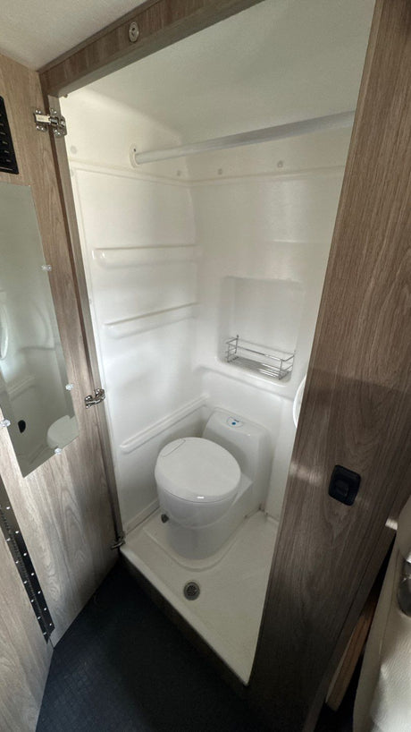 An RV rental's compact wet bath showing a white toilet, shower pan, and wall-mounted storage rack within the integrated shower space.