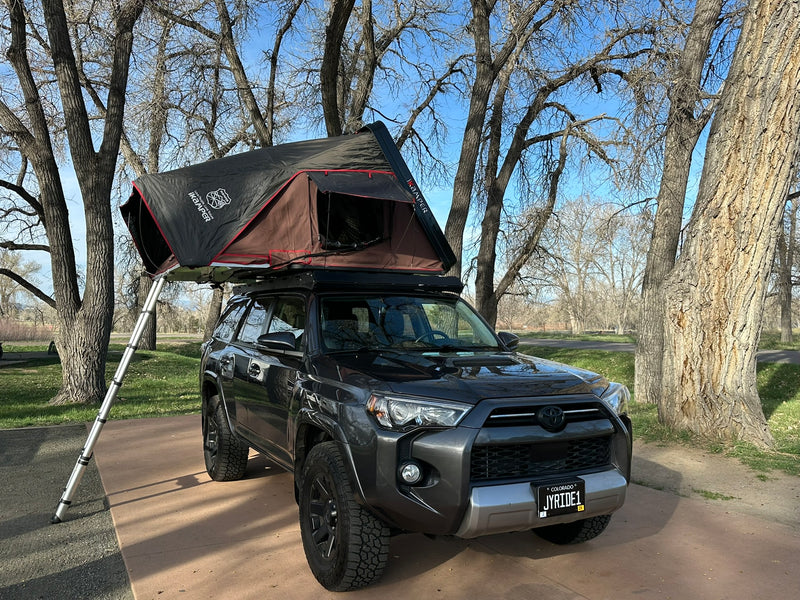 This dark gray SUV with a deployed iKamper rooftop tent offers a compact RV rental alternative for camping and outdoor adventures.|An RV rental SUV with a rooftop tent & ladder set up for camping — photo 1