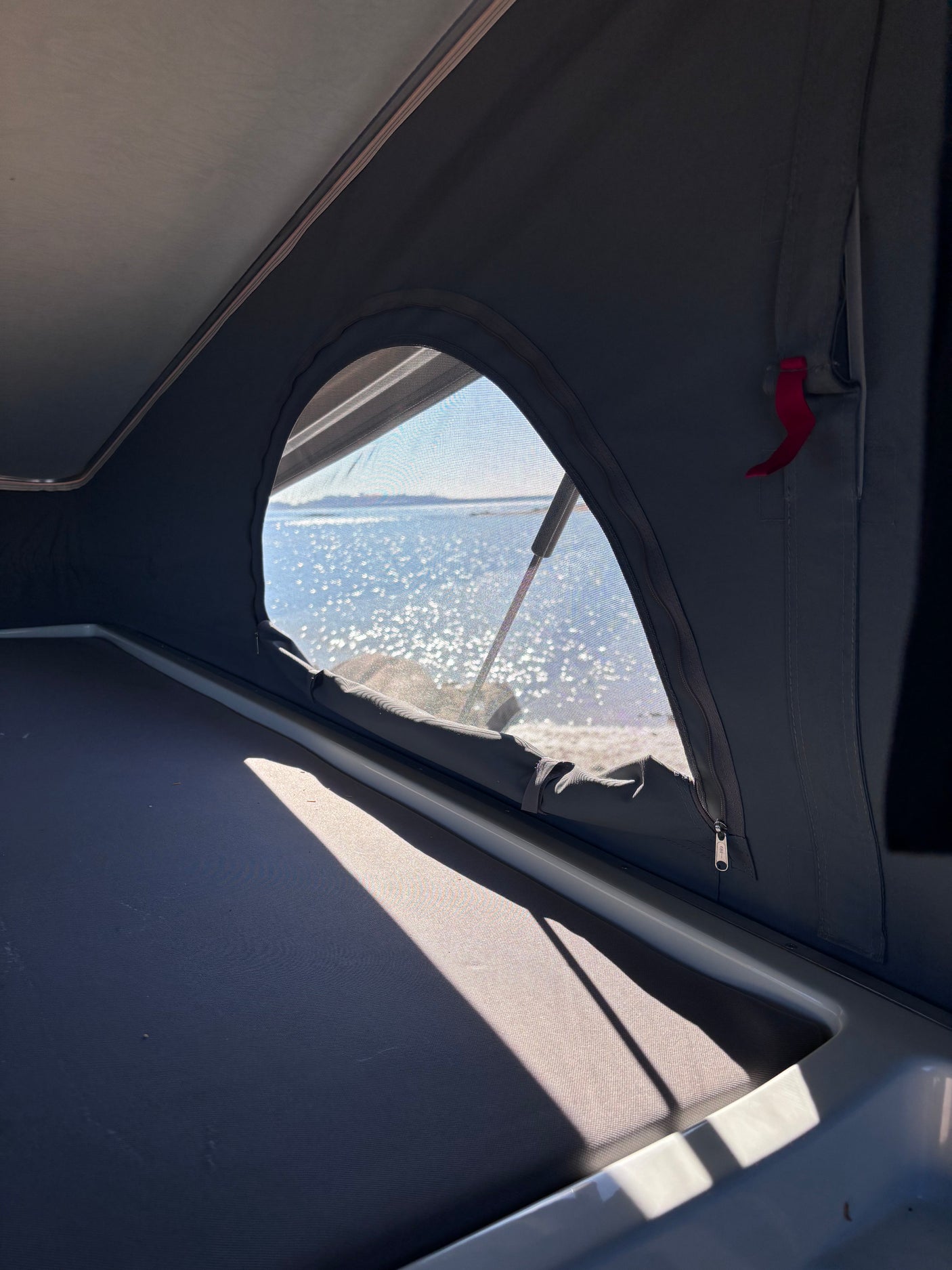 Inside an RV rental, a comfy bed faces a mesh window with sparkling water outside. Perfect for camping.