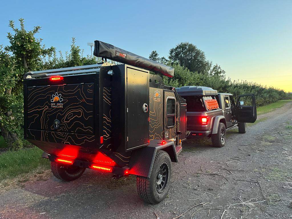  set against a stunning mountain backdrop.|Adventure-ready RV rental: This black off-road camping trailer with topographic graphics is hitched to a Jeep Gladiator truck.|An RV rental overlanding adventure at a forest campsite. A man relaxes beside a rugged trailer & Jeep with a rooftop tent.|Cozy forest RV rental campsite: black adventure vehicle with open kitchen